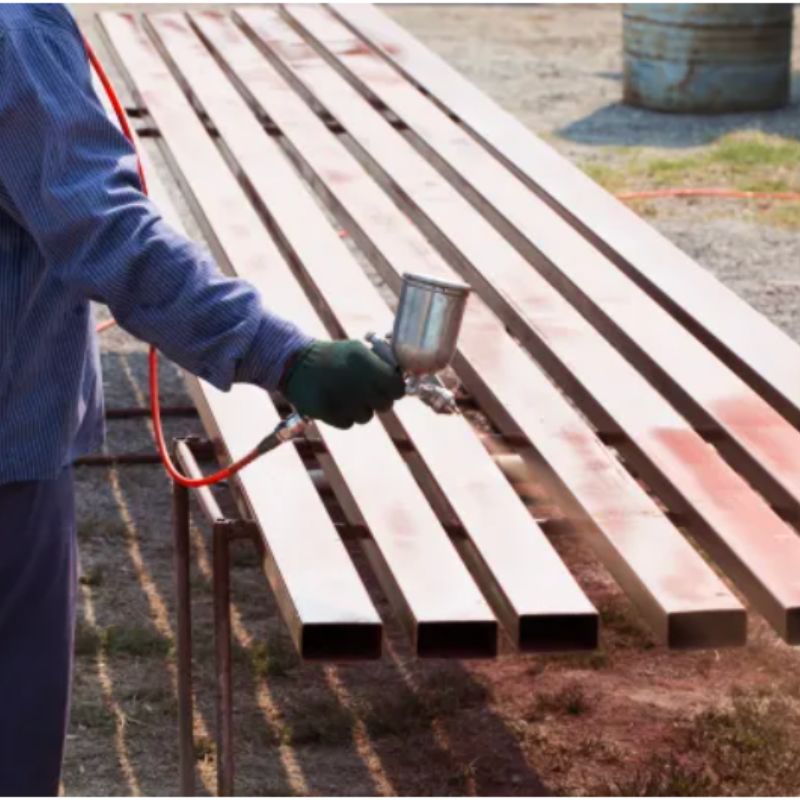 Worker applying Cortec VpCI-391 anti rust non tacky coating with a spray tool on metal bars placed outdoors.