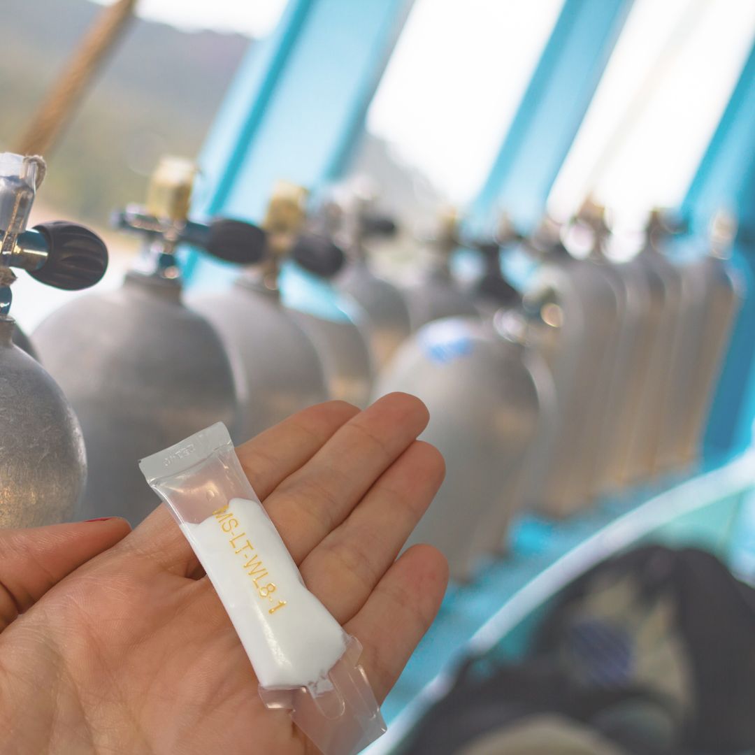 Hand holding a small white tube of Christo-Lube MCG 111 in front of oxygen cylinders.