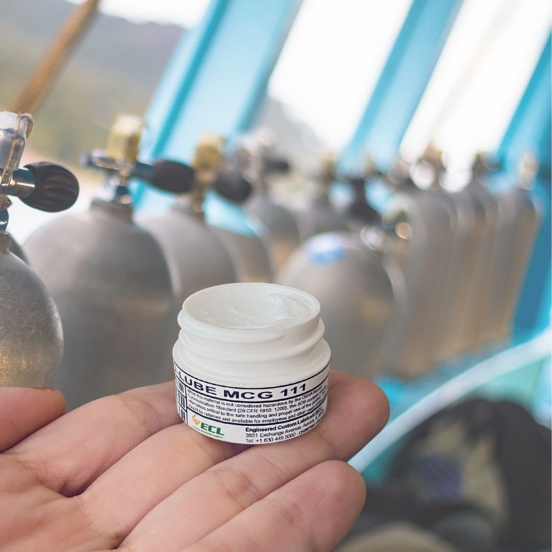 Hand holding a small jar of Christo Lube MCF 111 in front of oxygen cylinders. 
