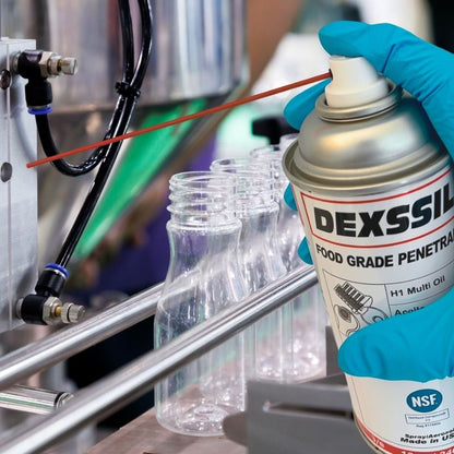 A hand wearing a blue nitrile glove holds a can of DEXSSIL Food Grade Penetrant spray near a metal production line with clear plastic bottles.